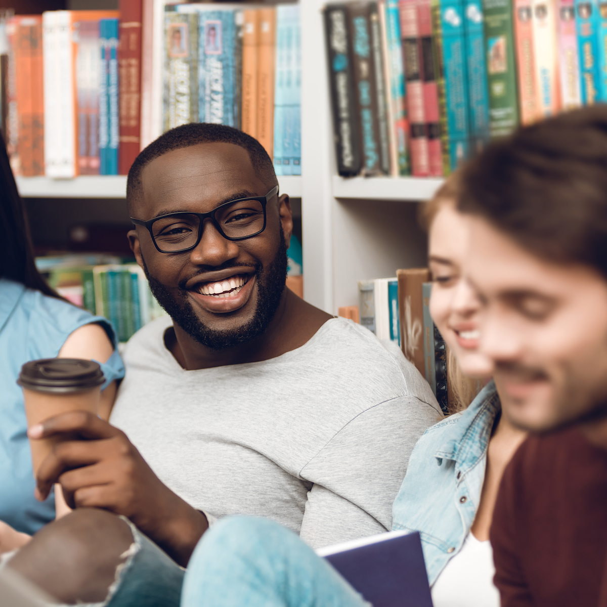 Group of ethnic multicultural students sitting, smiling and talking near bookshelf in library.